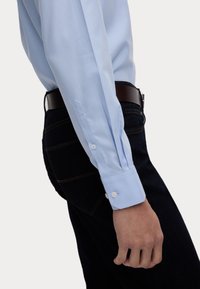 Ljusblå långärmad skjorta med knappar och spetsiga manschetter, gjord av mjukt tyg. Sitter bekvämt över mörka jeans med ett brunt bälte.