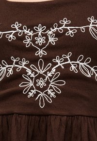 Tissu marron avec des motifs floraux et de feuilles blancs brodés formant des dessins décoratifs symétriques.