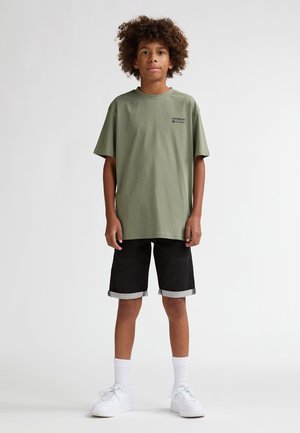 Camiseta de algodón verde oliva con un pequeño logo negro, combinada con shorts de mezclilla negros con puños doblados y zapatillas blancas.