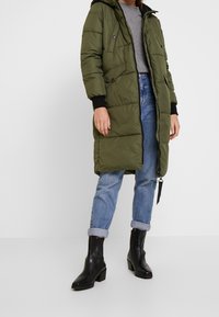 Oliwkowa kurtka puchowa z kapturem, z zamkiem błyskawicznym, przednimi kieszeniami i czarnymi mankietami. Noszona na jasnoniebieskie dżinsy i czarne botki na obcasie.