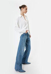 Weißes Hemd mit langen Ärmeln, kombiniert mit hellblauen weit geschnittenen Jeans. Das Outfit zeichnet sich durch einen lässigen Schnitt und ein minimalistisches Design aus.