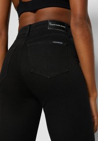 Persona vistiendo jeans de tiro alto negros de Calvin Klein, mostrada desde la parte trasera de la cintura hasta la mitad del muslo, con parches de marca en la cinturilla y el bolsillo.