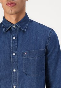 Mørkeblå denimskjorte med knapper foran, spiss krage, brystlomme og liten logodetalj over lommen. Jevn tekstur.