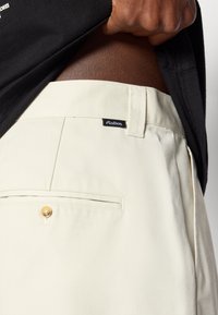 Μπεζ παντελόνι από υφασμάτινο υλικό με υφή, διαθέτοντας μία εμπρόσθια τσέπη με χρυσό κουμπί και μια μαύρη ταμπέλα μάρκας στο λάστιχο της μέσης.