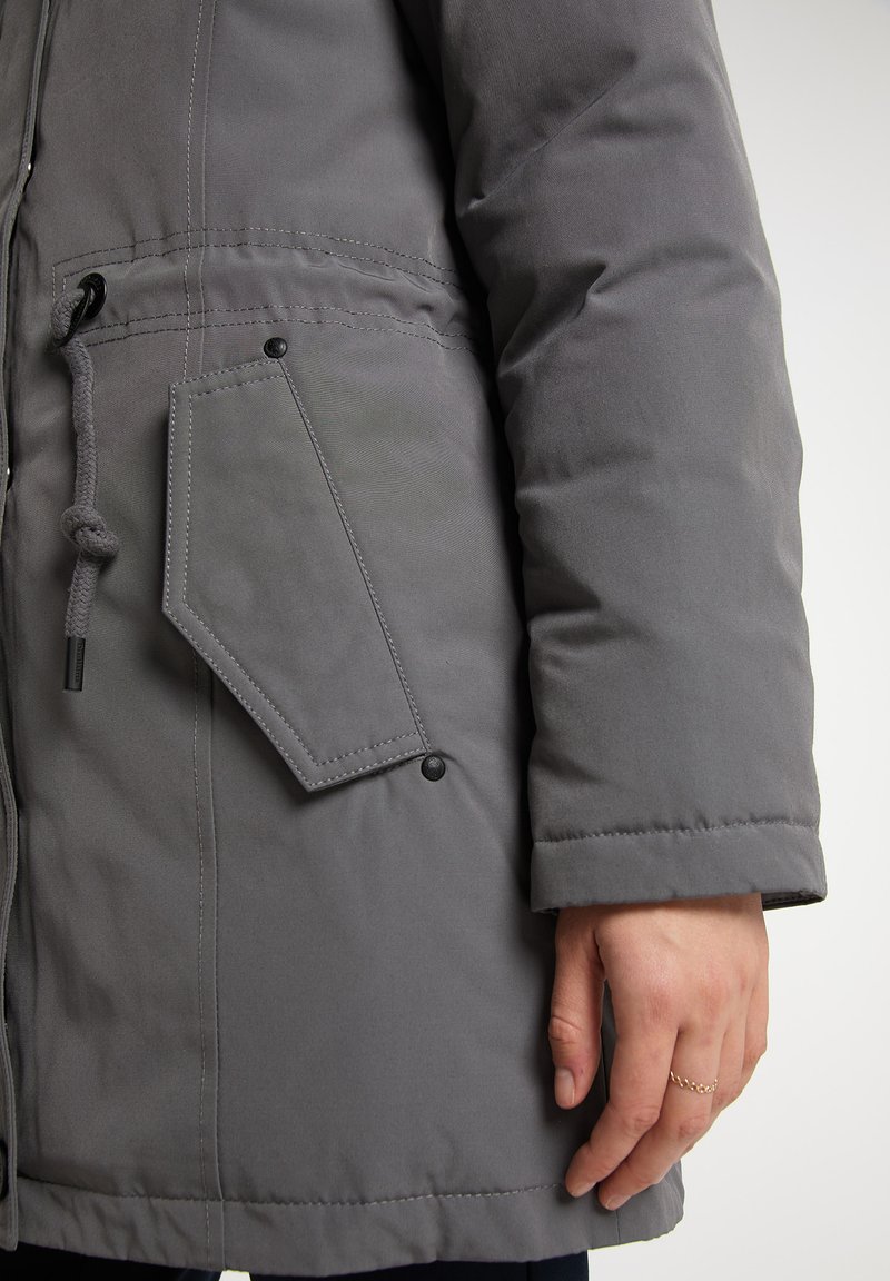 Graue gepolsterte Jacke mit einer Fronttasche und geometrischer Klappe, Kordelzug in der Taille und sichtbaren Nähdetails.