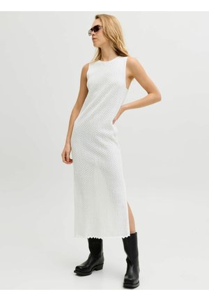 Mujer con vestido midi blanco sin mangas y textura, botas negras hasta la mitad de la pantorrilla y gafas de sol, de pie con una mano en la cadera sobre fondo liso.