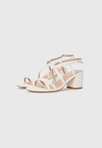 White heeled sandals with multiple crossed straps, a block heel, and an ankle strap, featuring a smooth texture and a minimalist design.