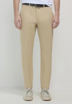 Beige Chinos mit geradem Schnitt, leichtem Stoff, Gürtelschlaufen und einer glatten Textur. Kombiniert mit hellgrauen Schuhen für einen lässigen Look.