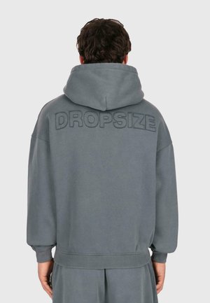 Mann in einem lockeren grauen Kapuzenpullover mit "DROPSIZE" auf dem oberen Rücken gestickt, der vor einem schlichten hellen Hintergrund steht.