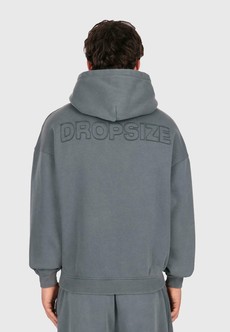 Mann in einem lockeren grauen Kapuzenpullover mit "DROPSIZE" auf dem oberen Rücken gestickt, der vor einem schlichten hellen Hintergrund steht.