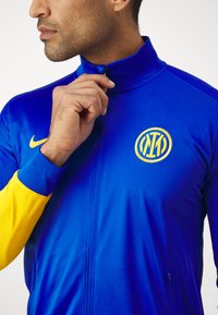 Giacca da ginnastica blu Nike con zip completa, dotata di maniche gialle e di un logo rotondo ricamato sul petto. Tessuto dalla texture liscia.