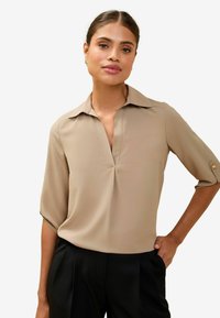 Blusa beige con cuello, escote en V y mangas tres cuartos. Presenta una textura suave y pliegues sutiles. Los pantalones negros completan el look.