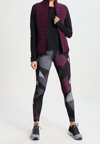 Donna che indossa una maglietta nera a maniche lunghe, un gilet trapuntato viola, leggings geometrici neri e grigi, e scarpe da ginnastica grigie in piedi.