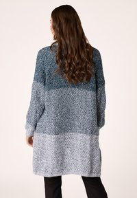 Cardigan oversize lavorato a maglia in tonalità di teal e grigio chiaro, con un motivo testurizzato e maniche lunghe. Orlo arrotondato e fessure laterali.