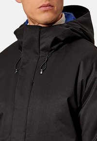 Giacca impermeabile nera con collo alto e cappuccio regolabile. Presenta una fodera blu a contrasto e chiusure frontali per fissare il cappotto.