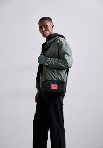 Grüne Bomberjacke mit gemustertem Ärmel, über einem schwarzen Hoodie getragen. Schwarze Umhängetasche mit rotem Label und komplizierten Mustern. Schwarze Hose.