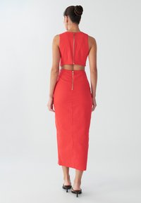 Crveni dvodijelni komplet koji se sastoji od kratkog topa s patentnim zatvaračem na leđima i suknje visokog struka do gležnjeva s odgovarajućim patentnim zatvaračem. Glatki, teksturirani materijal.