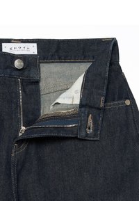 Jean en denim bleu foncé avec fermeture éclair et bouton ouverts, montrant le tissu de la poche intérieure et une étiquette blanche avec texte japonais et site web.