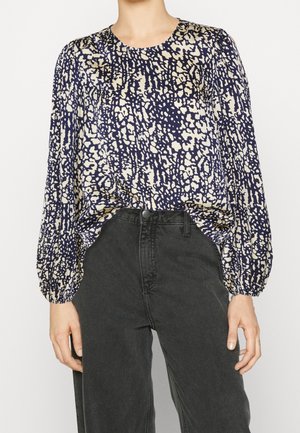 Blusa blu navale con un motivo astratto beige, scollatura rotonda e maniche lunghe a sbuffo, abbinata a jeans neri a vita alta. Tessuto morbido.
