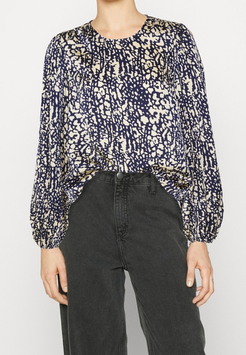 Blusa blu navale con un motivo astratto beige, scollatura rotonda e maniche lunghe a sbuffo, abbinata a jeans neri a vita alta. Tessuto morbido.