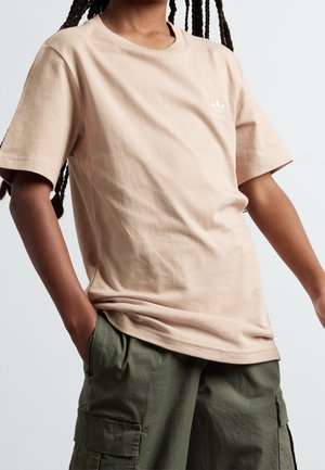 Person med flettet hår iført en beige Adidas t-shirt og grønne cargobukser, hænderne i lommerne, foran en ensfarvet lys baggrund.