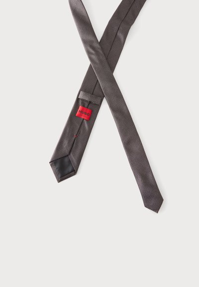 Cravate texturée gris foncé avec une petite étiquette rouge de marque et un détail noir à la pointe, posée en croix sur fond blanc.