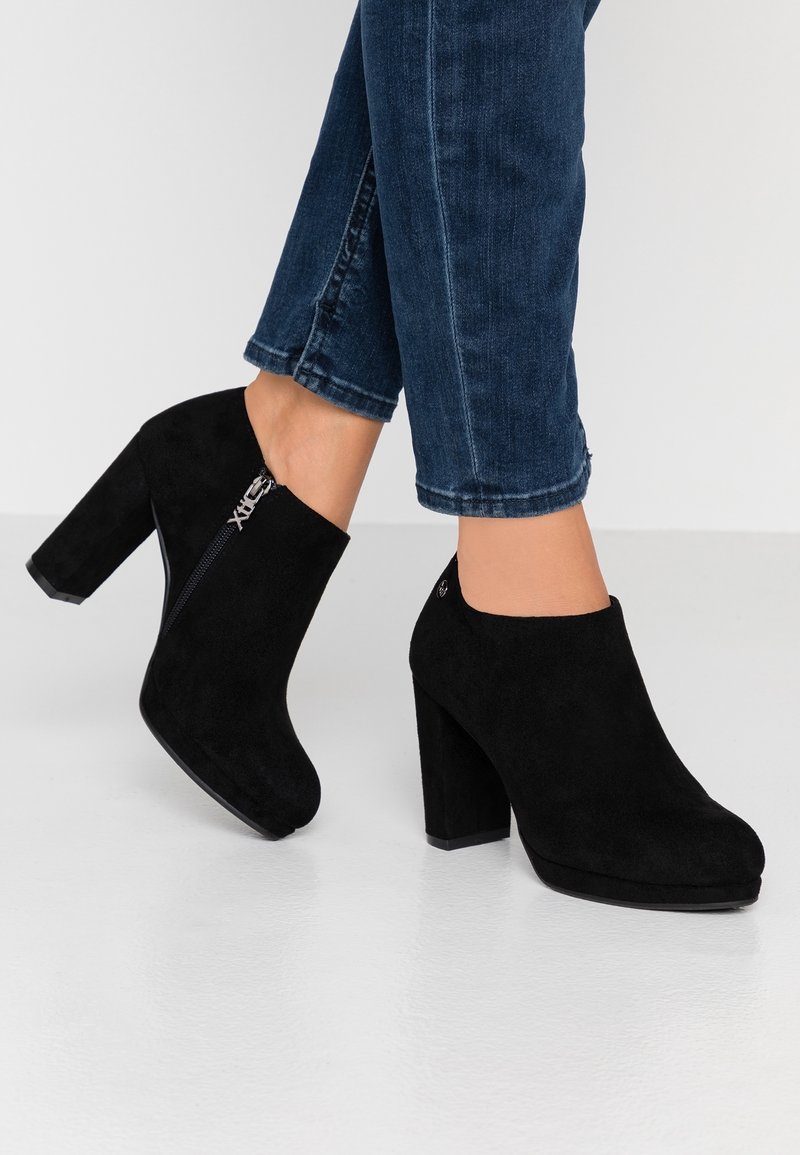 Bottines en daim noir avec talons carrés et fermetures éclair latérales, portées avec un jean en denim bleu foncé sur un sol blanc.