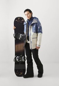 Fekete és lila árnyalatú snowboard, rajta rögzített kötésekkel; személy kékfehér virágos dzsekiben, fekete nadrágban és csizmában.