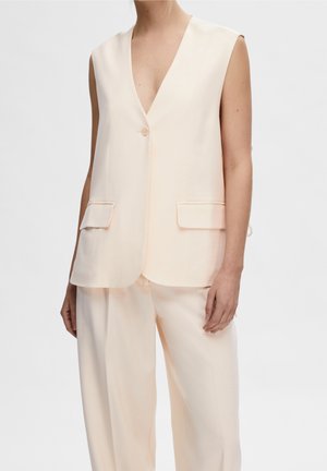 Femme portant un gilet crème sans manches avec un bouton et un pantalon large assorti sur un fond uni.