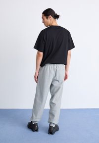 Schwarzes Baumwoll-T-Shirt kombiniert mit hellgrauen Jogginghosen. Die Hose hat einen elastischen Bund und eine Gesäßtasche. Die Schuhe sind schwarze Sportschuhe.