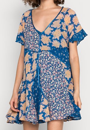 Niebieska sukienka z mieszanymi kwiatowymi wzorami w kolorze brzoskwiniowym i białym, z dekoltem w serek i krótkimi falbanianymi rękawami, noszona przez stojącą osobę.