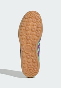 Gummisula i tan med cirkulära mönster och vågmönster. Sidan har gröna och lila detaljer med logotyp.