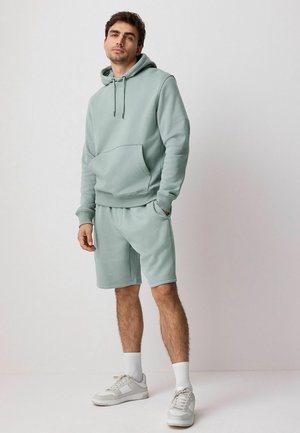 Felpa verde chiaro con grande tasca frontale e lacci, abbinata a pantaloni corti coordinati. Sneakers e calze bianche completano l'outfit.
