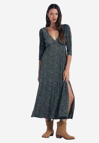 Grünes Wickelkleid mit Muster, 3/4-Ärmeln und seitlichem Schlitz, kombiniert mit braunen Stiefeletten. Der Stoff hat einen leichten Glanz und eine strukturierte Oberfläche.