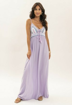 Femme aux longs cheveux bruns portant une robe maxi lilas sans manches avec un haut multicolore en crochet et des sandales, debout devant un fond blanc.