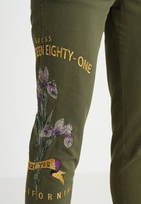 Pantalon vert olive avec des fleurs violettes brodées, texte doré indiquant "GUESS NINETEEN EIGHTY-ONE" et un ruban disant "LUCKY YOU" au-dessus de "CALIFORNIA."