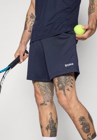 Marineblaue Sportshorts mit weißem "BORG"-Logo, kombiniert mit einem passenden kurzärmeligen Shirt, hält einen Tennisschläger und einen Ball.