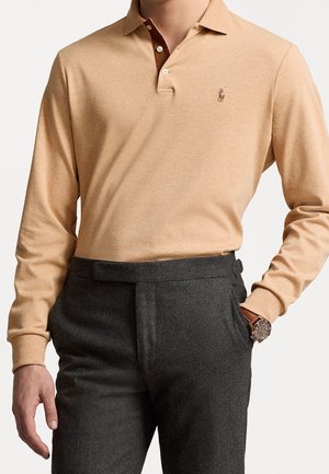 Homme portant un polo à manches longues beige avec un petit logo brodé, un pantalon gris foncé, et une montre en cuir marron au poignet gauche.