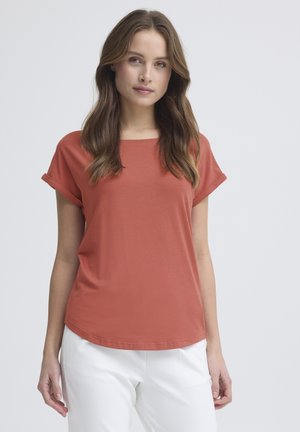 Vrouw met lang bruin haar, gekleed in een roestkleurige korte mouw shirt en witte broek, staande tegen een lichtgrijze achtergrond.