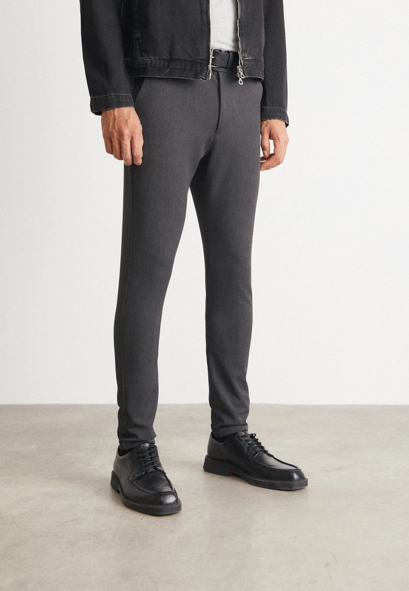 Pantalons gris fuselés en tissu extensible, avec une texture lisse. Associés à des chaussures noires à lacets et une veste en denim noire.