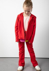 Czerwona bluza rozpinana, miękki materiał, dwie przednie kieszenie, haftowane detale. Połączona z czerwonymi spodniami oraz warstwowymi topami w kolorze różowym i żółtym. Białe sneakersy.