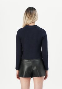 Vrouw met lang blond ombre-haar in een zwart langslavende top en zwarte leren shorts, staand met de rug naar de camera tegen een witte achtergrond.