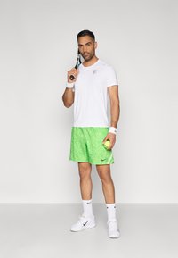 Λευκό αθλητικό μπλουζάκι, πράσινα κοντά σορτς με πουά σχέδιο, κρατώντας μια ρακέτα τένις και μια μπάλα. Λευκά αθλητικά παπούτσια και κάλτσες με μαύρες λεπτομέρειες.