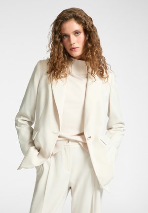 Frau mit lockigem braunem Haar, die ein cremefarbenes Blazer, passende Hosen und einen hochgeschlossenem Pullover trägt, steht mit den Händen in den Taschen.