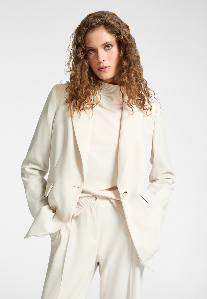 Mujer con cabello rizado castaño, vestida con un blazer blanco roto, pantalones a juego y un suéter de cuello alto, de pie con las manos en los bolsillos.