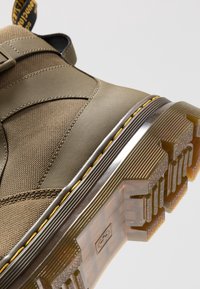 Gros plan d'une chaussure en tissu beige texturé avec des coutures jaunes et une semelle épaisse en caoutchouc translucide montrant des motifs de la bande de roulement.