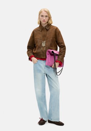 Młoda kobieta o krótkich blond włosach, ubrana w brązową kurtkę, czerwony sweter, jasnoniebieskie dżinsy, czarne buty i naszyjnik, trzyma różową torebkę na wysokości talii.