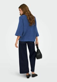 Femme aux cheveux longs et ondulés portant un pull bleu, un jean large foncé, des ballerines noires, tenant un sac à main noir, debout devant un fond clair.