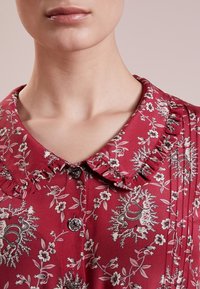 Czerwona bluzka w kwiaty z falbaniastym kołnierzykiem i detalem guzika. Posiada skomplikowane czarno-białe wzory botaniczne oraz lekką, teksturowaną tkaninę.