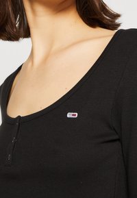 Schwarzes, geripptes Langarmshirt mit Rundhalsausschnitt und Knopfdetail, ausgestattet mit einem kleinen gestickten Logo auf der linken Brust.